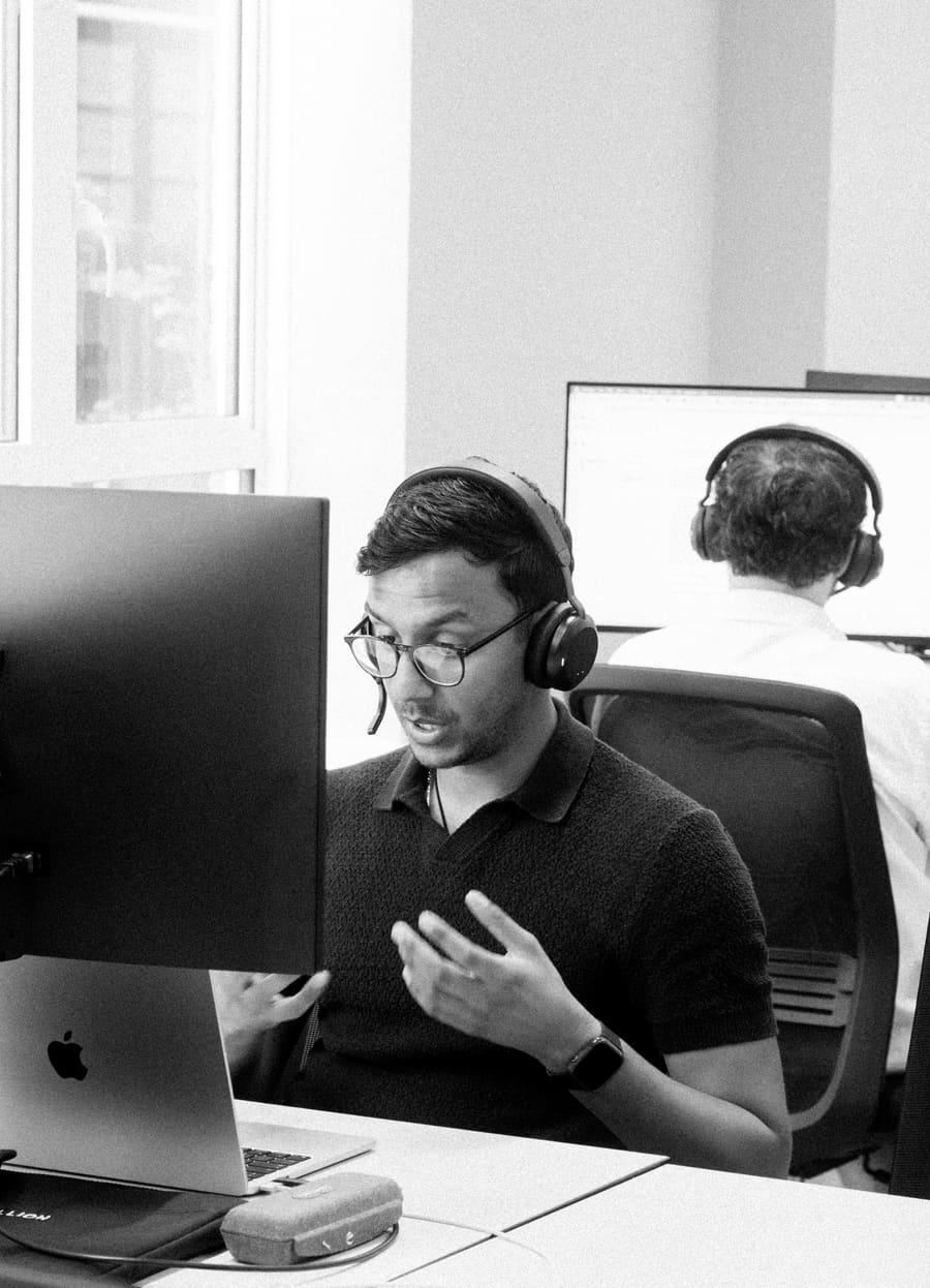 A teammate working at his desk with headphones on, a colleague behind him